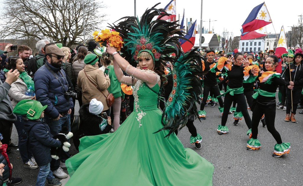 Photo of the Philippines performance for the 2023 St. Patrick's Parade in Galway. A woman in a green dress with a feathered head crown is in a dancing pose with children behind her holding their flag and following her 