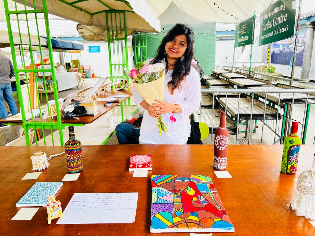 Namrata stands behind her stall, which features several colourful works of art - including pottery and paintings. She is smiling and holding a bouquet of flowers.