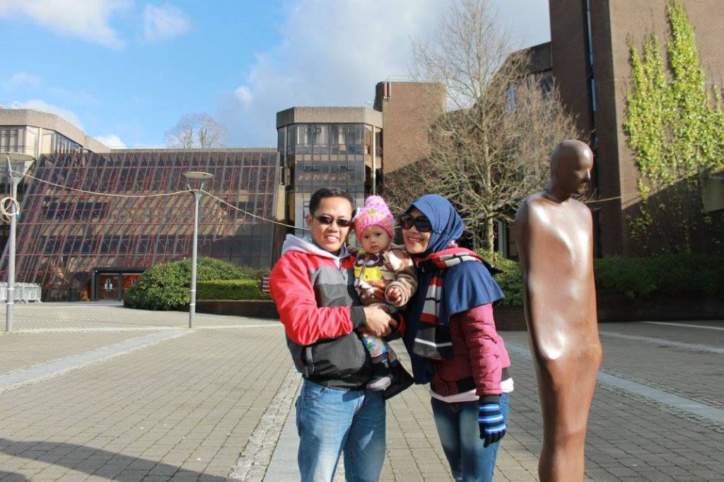 Yanuar stands beside Brown Thomas holding his baby with his wife standing beside him.

the main building is in the background