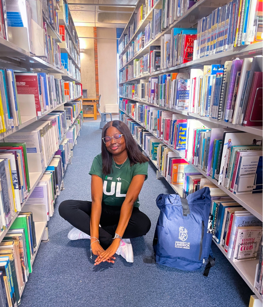 Augusta sitting on the blue carpet of the library floor in between shelves of books. She has her navy student ambassador school bag on the floor beside her