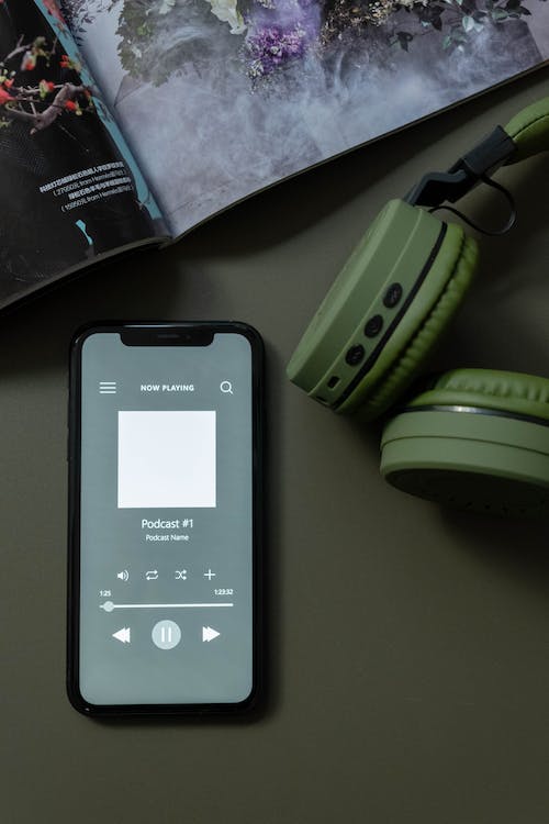 A phone with a podcast playng on it on the desk beside a pair of green headphones