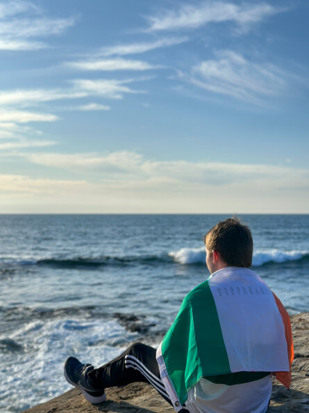 Daniel sitting at La Jolla Cove, California, watching the Pacific Ocean waves with an Irish flag wrapped around him.