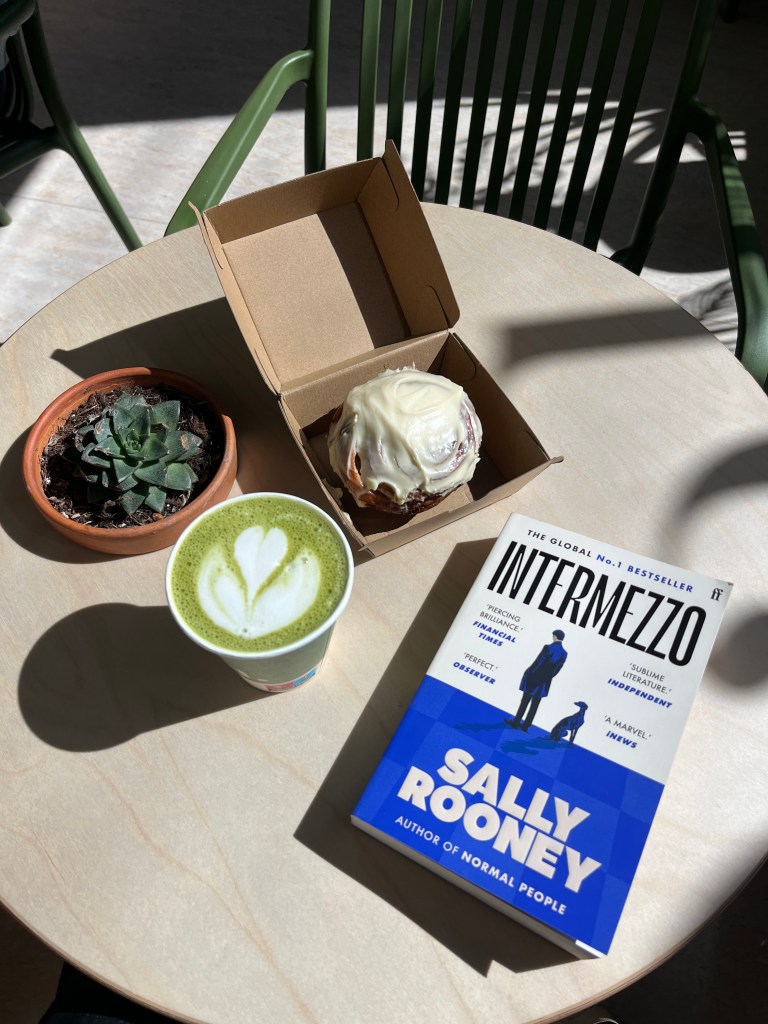 A matcha, cinnamon bun and the book Intermezzo by Sally Rooney arranged on a table