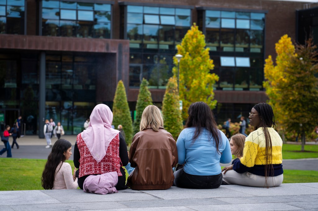 4 students sitting on the student centre steps facing away from the camera towards the Glucksman Library which is partially in the background