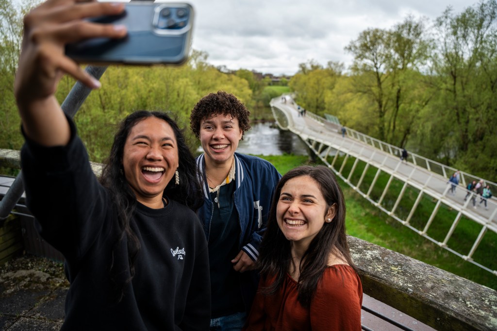 3 students take a selfie in a balcony with the Living Bridge behind them and lush greenery